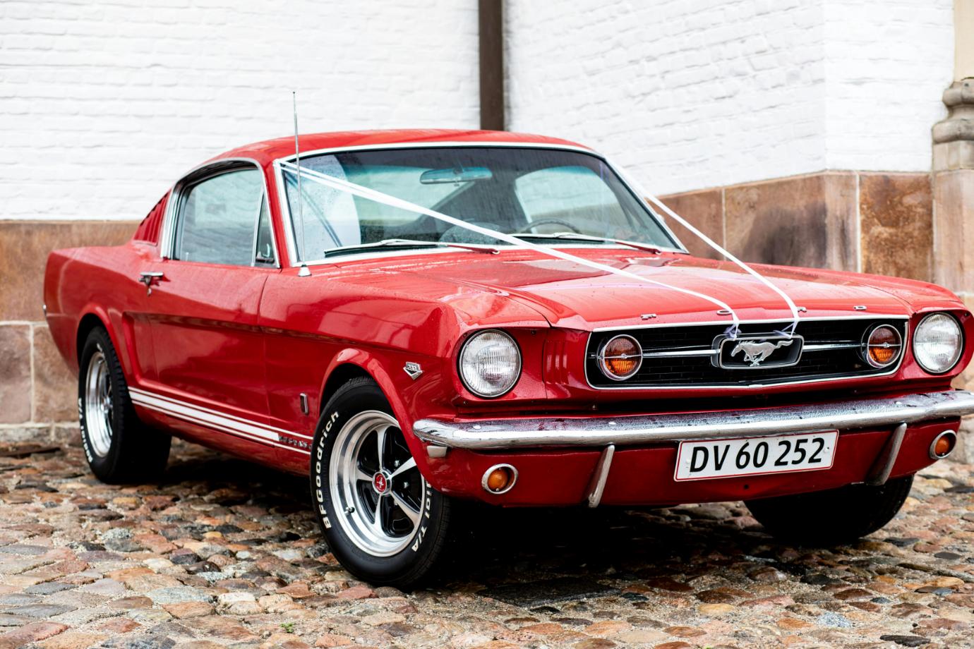 Kvinna i 60-talsinspirerad outfit med solglasögon och hårband vid en vit veteranbil på stadsgata