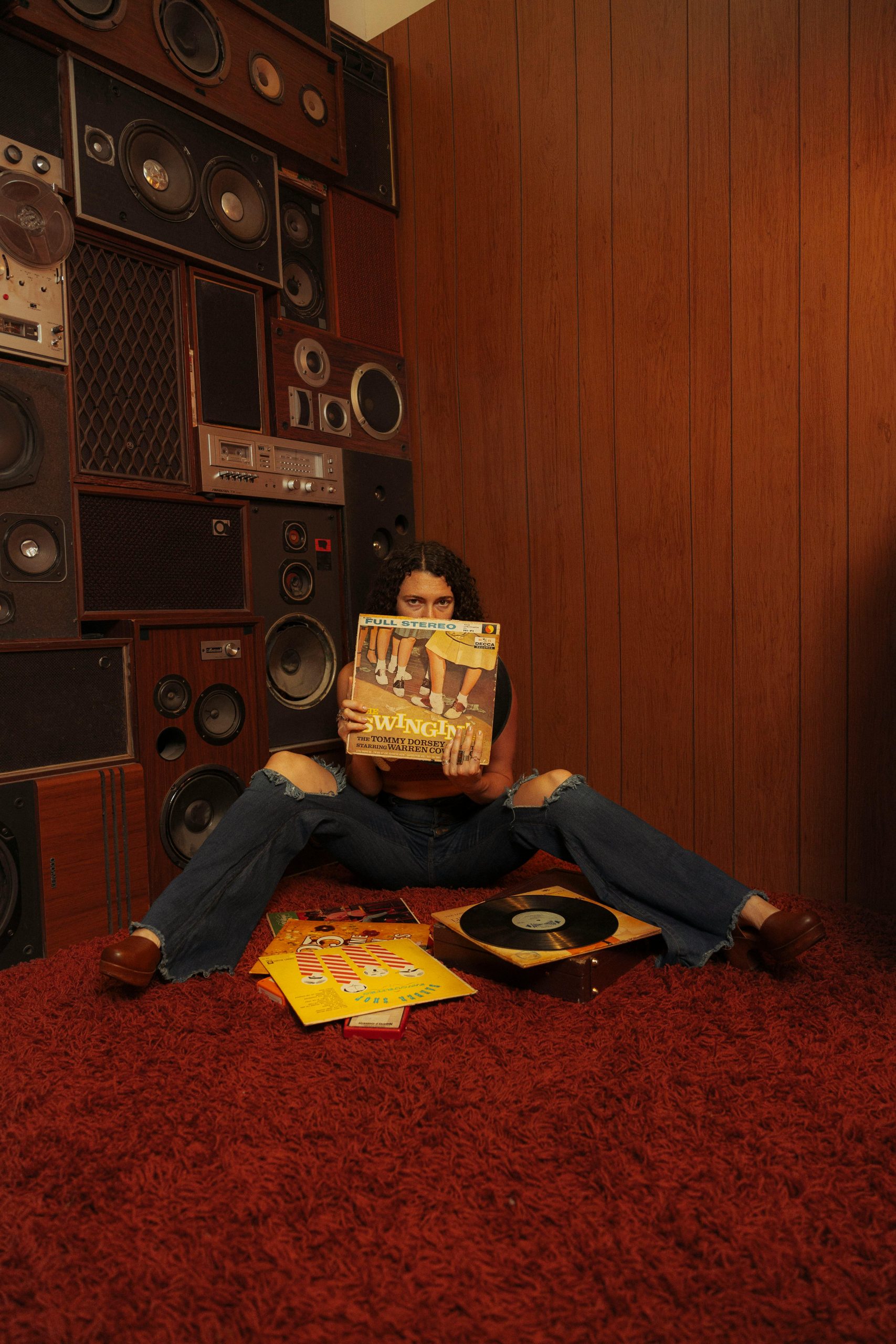 Person på röd matta framför en vägg av högtalare håller upp ett vinylomslag med fler skivor på golvet.